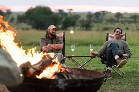 Two friends with sundowners beside a fire pit, looking out over the plains at dusk