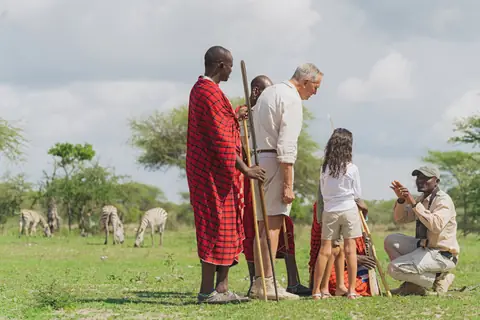 A family walking the grasslands with their Maasai guides, zebra grazing beyond