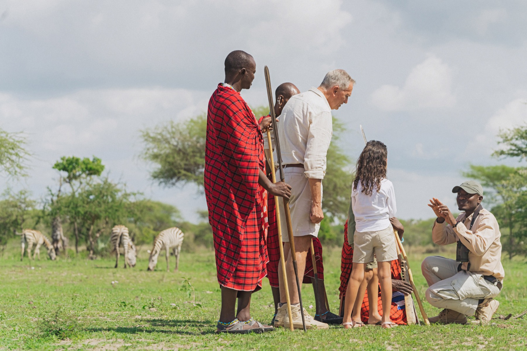 A grandfather and young girl on a walk with their Maasai guides, zebra grazing beyond